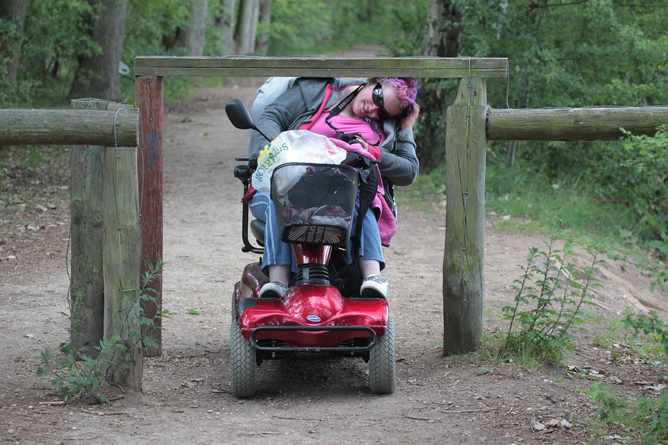 Een vrouw in een elektrische rolstoel die haar hoofd sterk zijwaarts moet buigen om onder een barrière door te rijden