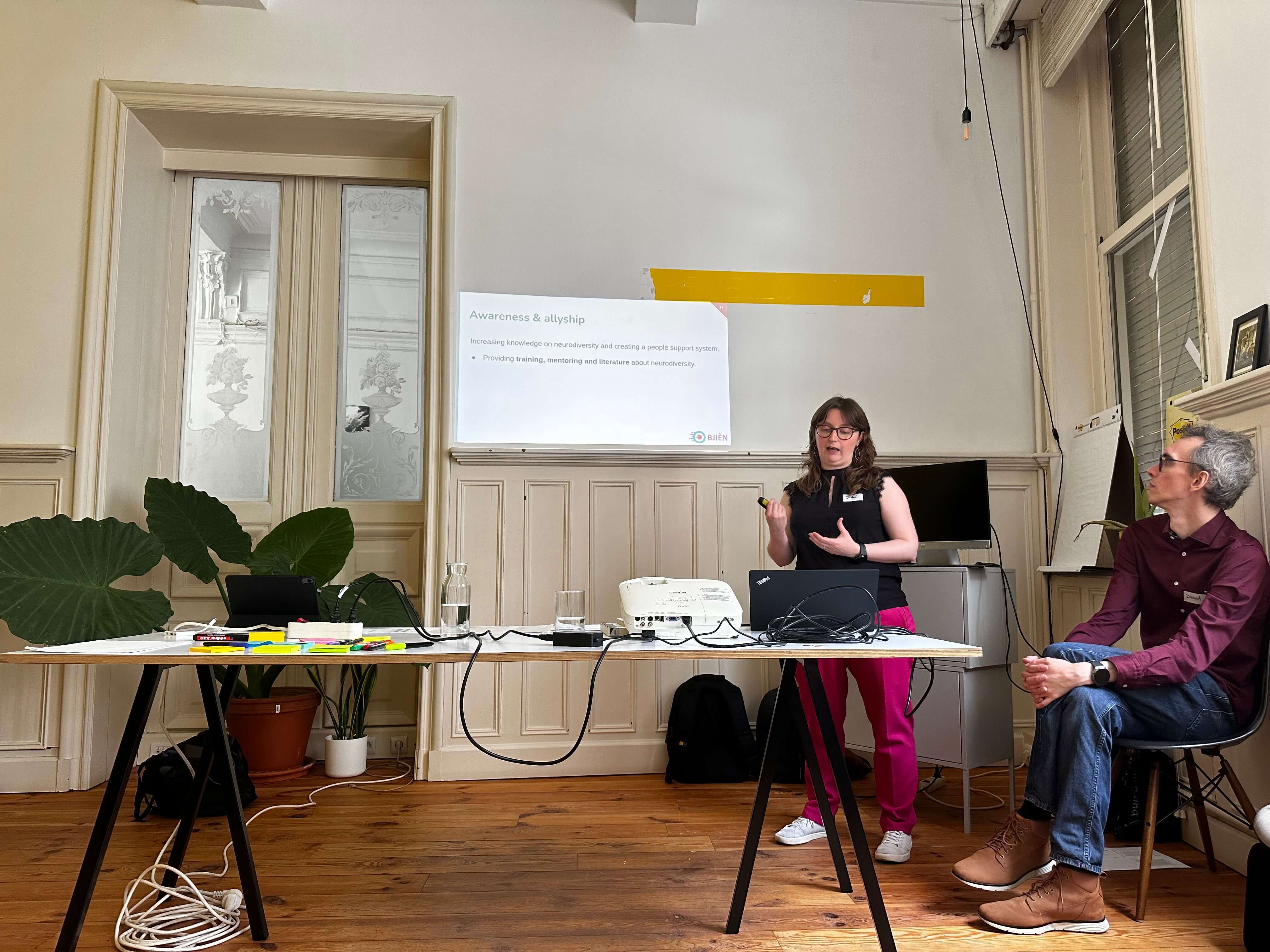 Daphné from Bjièn standing behind a table and presenting. Behind her, a slide with the title "Awareness & allyship" is visible. Her colleague Dietrich sits next to her and looks at the slide.